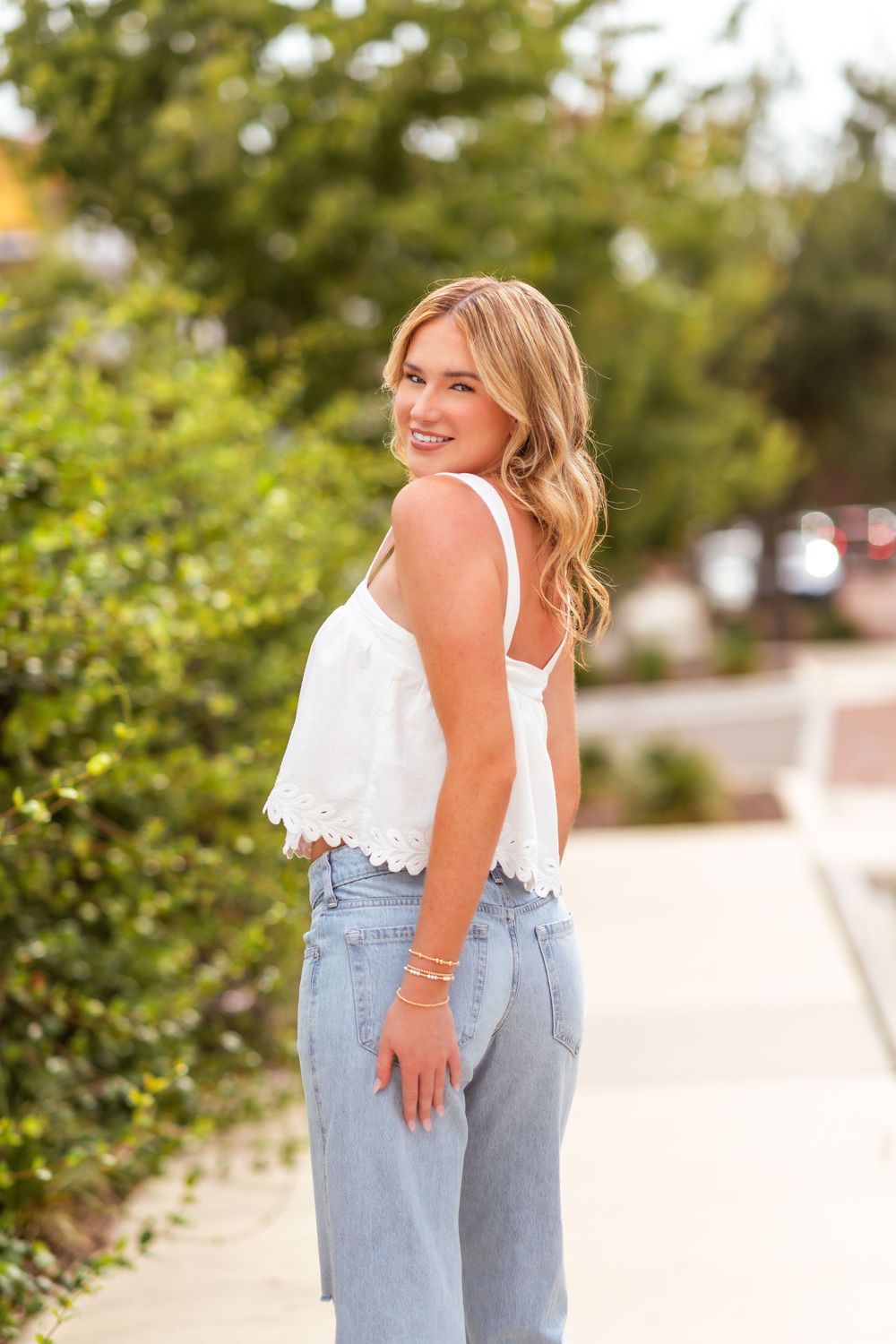 Young blonde woman in white crop top and light blue jeans smiling over shoulder outdoors.