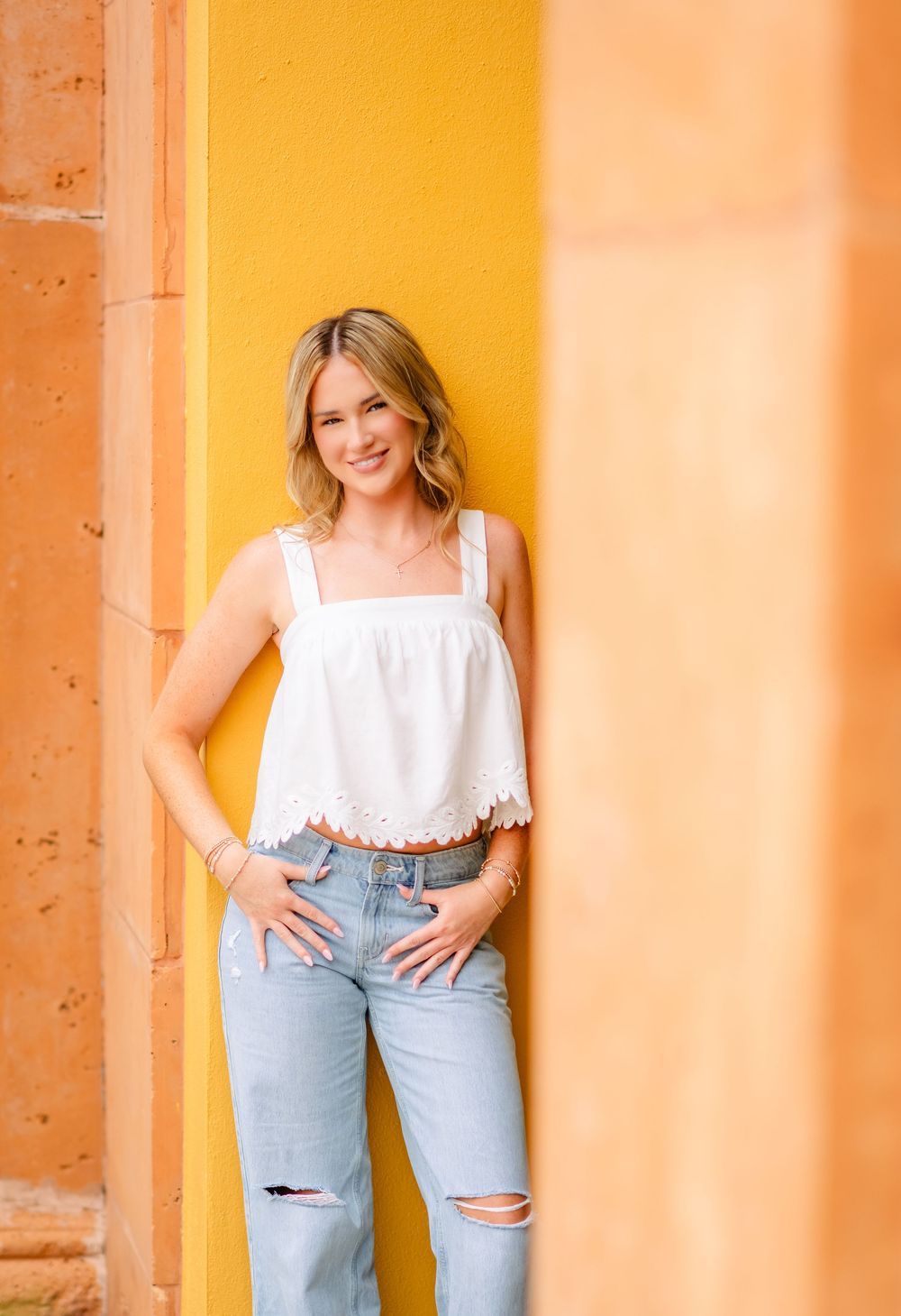 Young blonde woman smiling in white crop top and ripped jeans leaning against a bright yellow wall outdoors.
