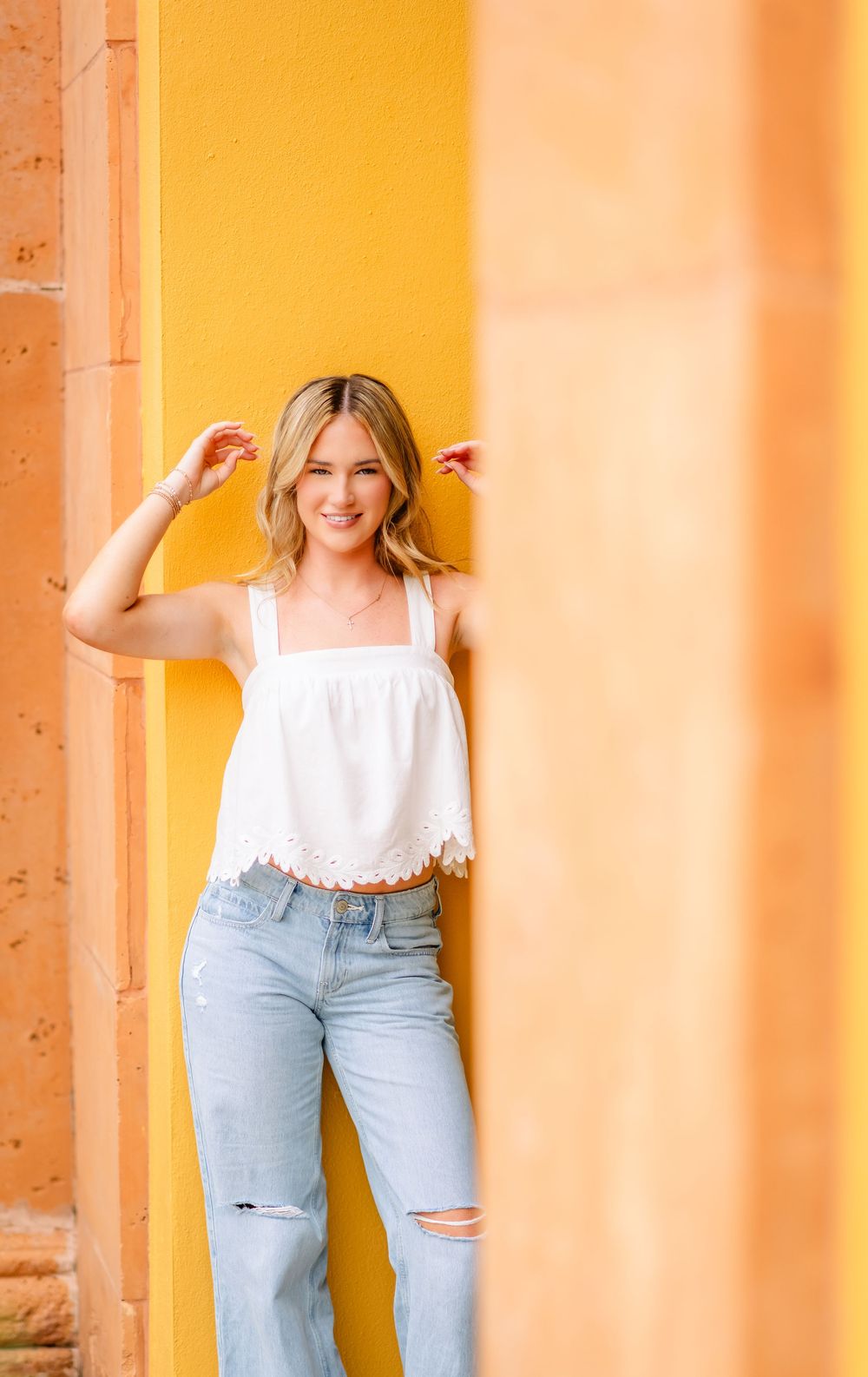 Smiling blonde woman in white crop top and ripped jeans leaning against a yellow wall.