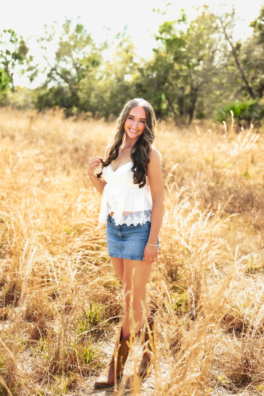 Young woman in white lace top and denim skirt smiling in golden field during senior portrait session.