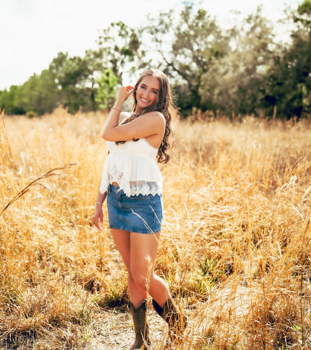 Young woman in white lace top, denim skirt, and cowboy boots posing in a golden sunlit field.