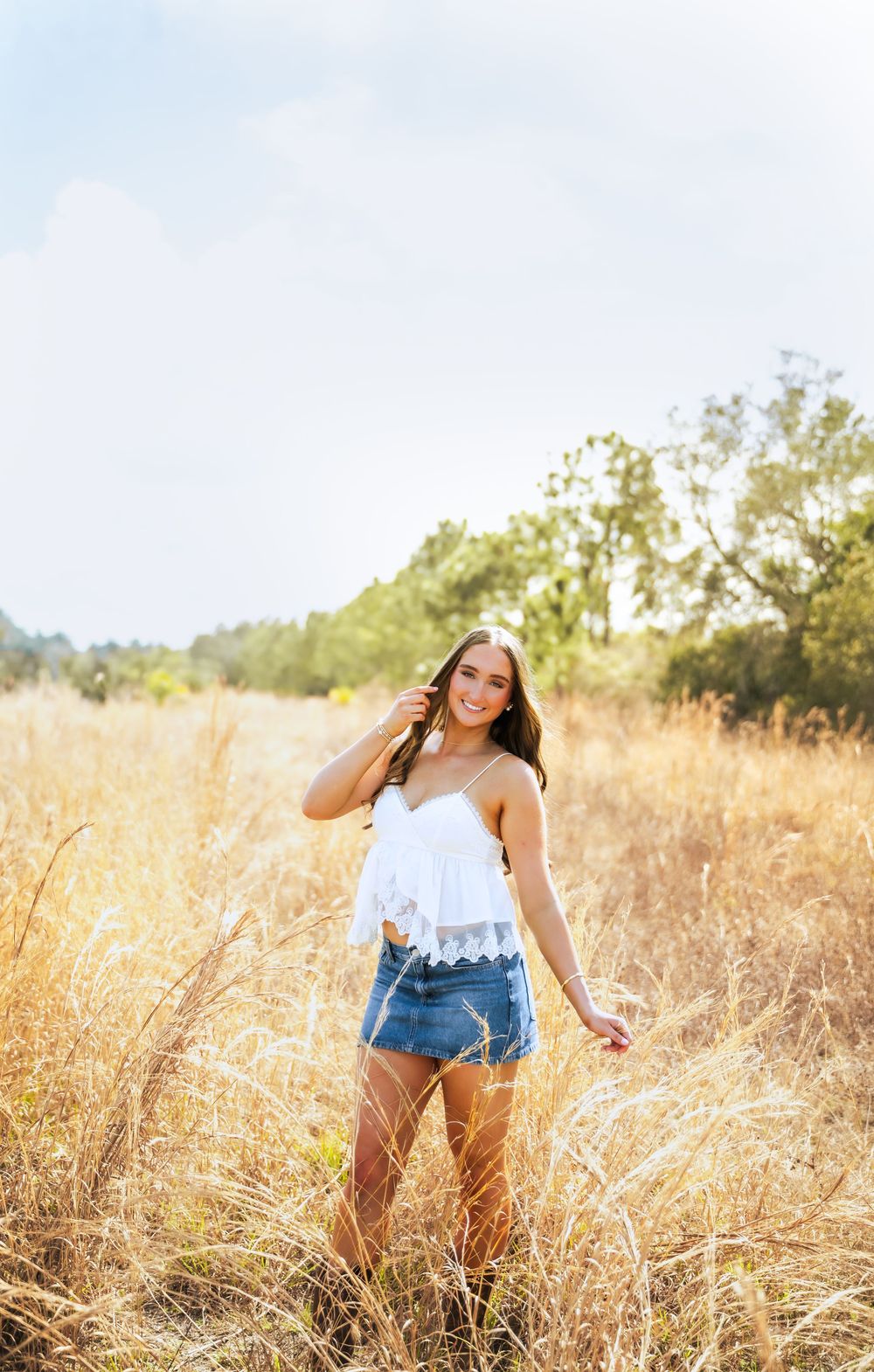 Young woman in white lace crop top and denim skirt smiling in golden field.