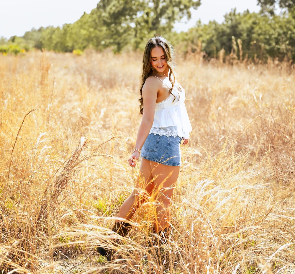 Young woman in white top and denim shorts walks through golden field with trees in background on sunny day.
