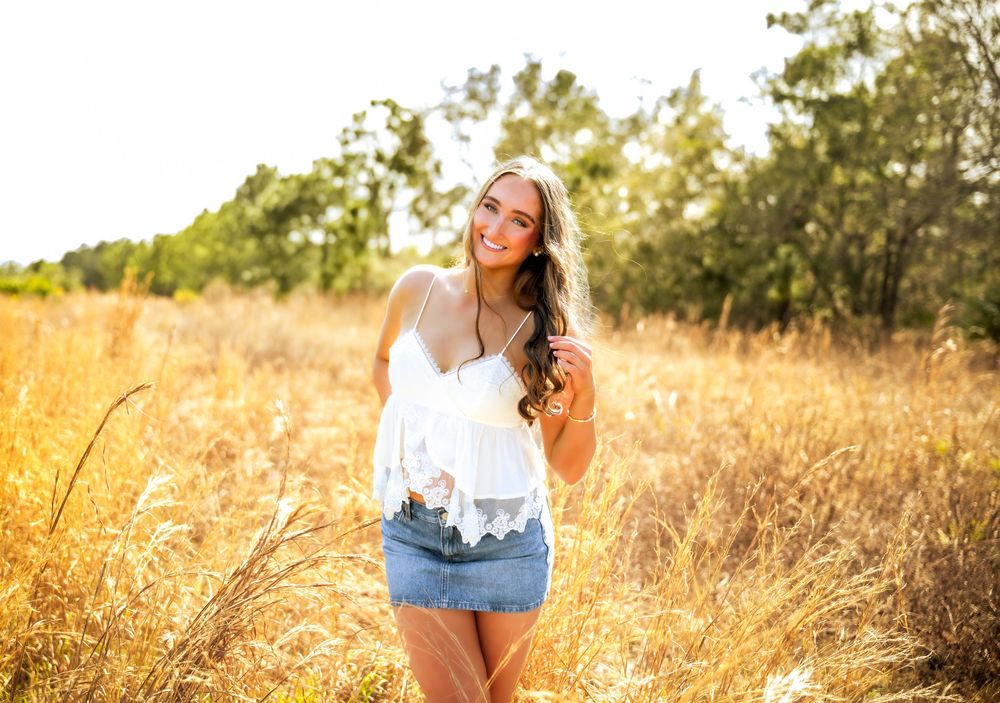 Young woman smiling in a golden field, wearing a white cami top and denim skirt on a sunny day.