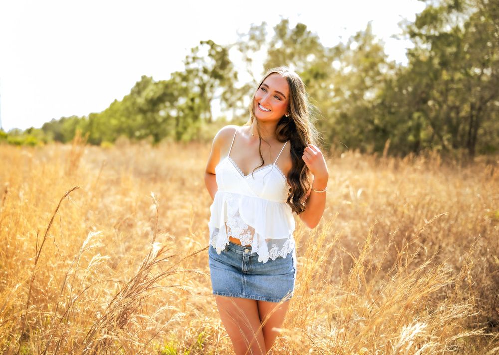Young woman in white lace top and denim shorts smiling in a golden field on a sunny day.