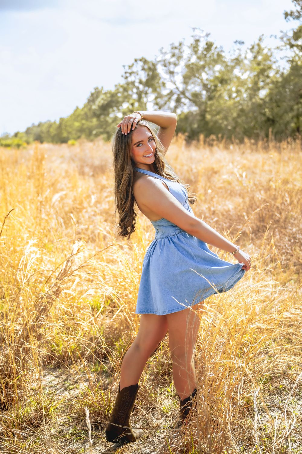 Smiling brunette woman in blue dress and cowboy boots twirling in sunny golden meadow.