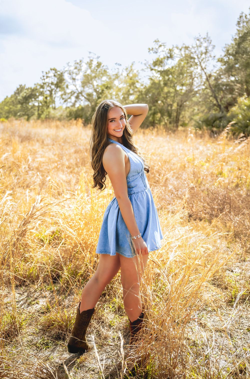 Young woman in blue dress and cowboy boots posing in golden field during sunny day.