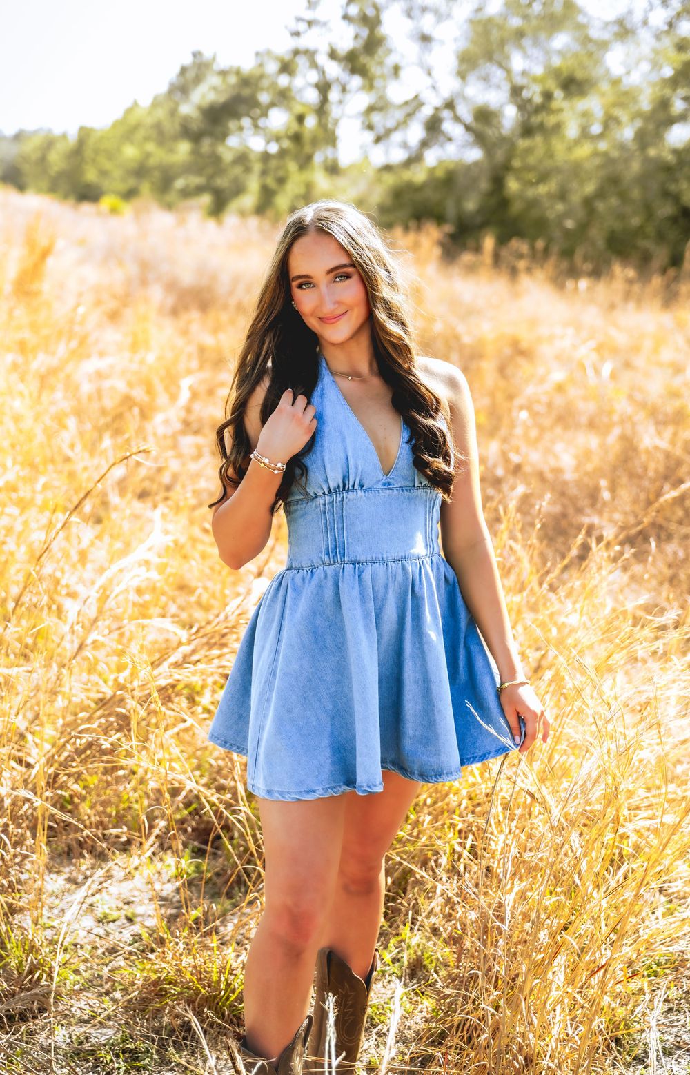 Young woman in blue denim dress and cowboy boots posing in golden field.