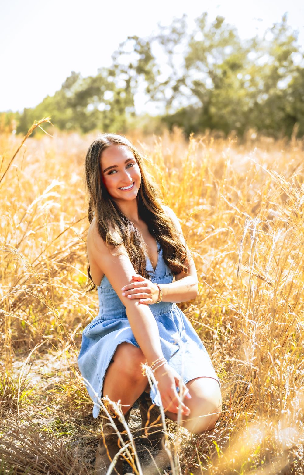 Smiling brunette teen in blue sundress kneeling in sunlit golden grass field for senior photos.