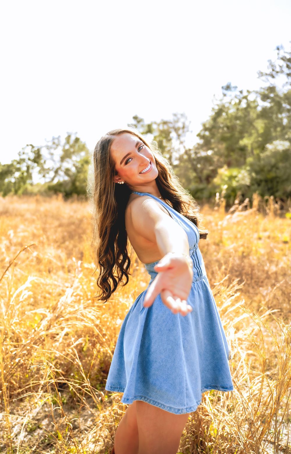 Smiling teen girl in blue denim dress reaching toward camera in golden field during sunny day.