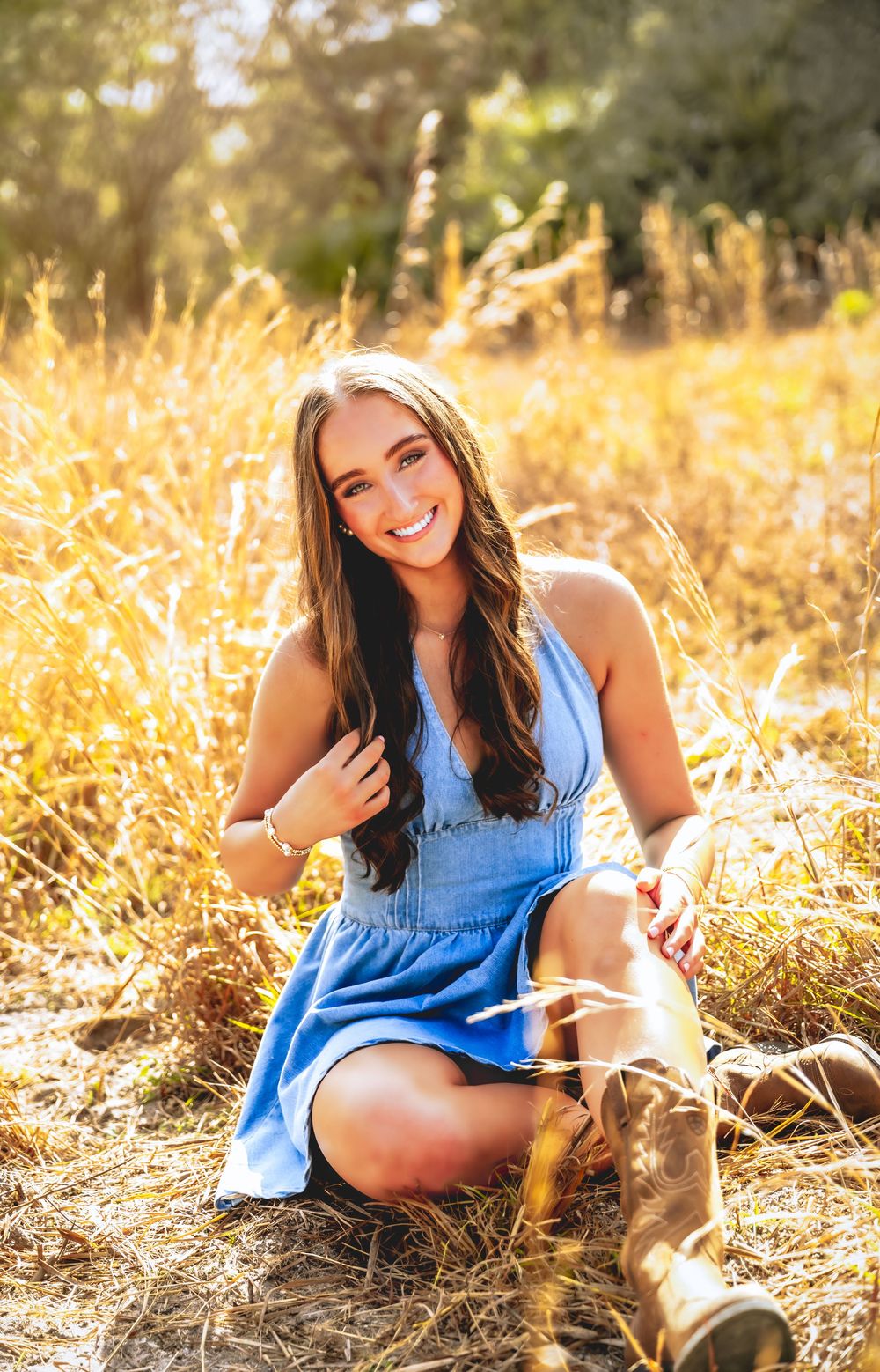 Smiling young woman in blue sundress and cowboy boots sitting in a sunlit golden grass field.