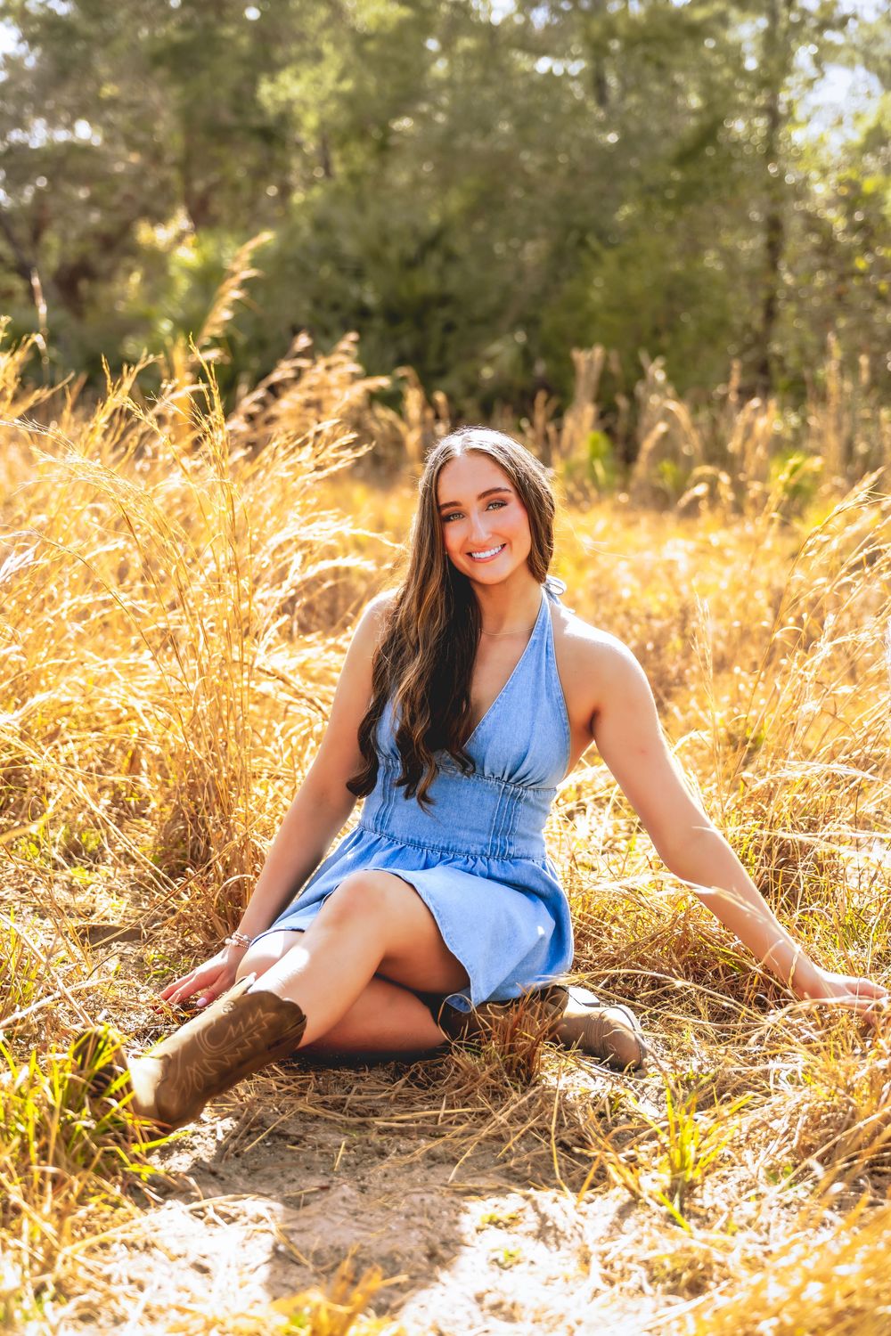 Young woman in blue denim dress and cowboy boots sits smiling in golden field of tall grass.