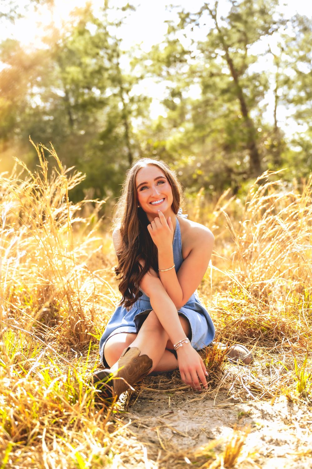 Young woman in denim romper and cowboy boots sitting in golden field grass with sunlit trees behind her.