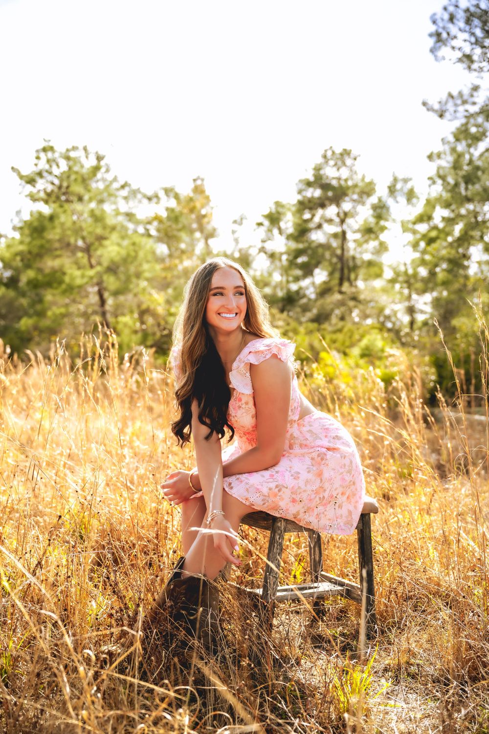 Smiling young woman in pink floral dress sits on stool in golden sunlit field with trees behind her.