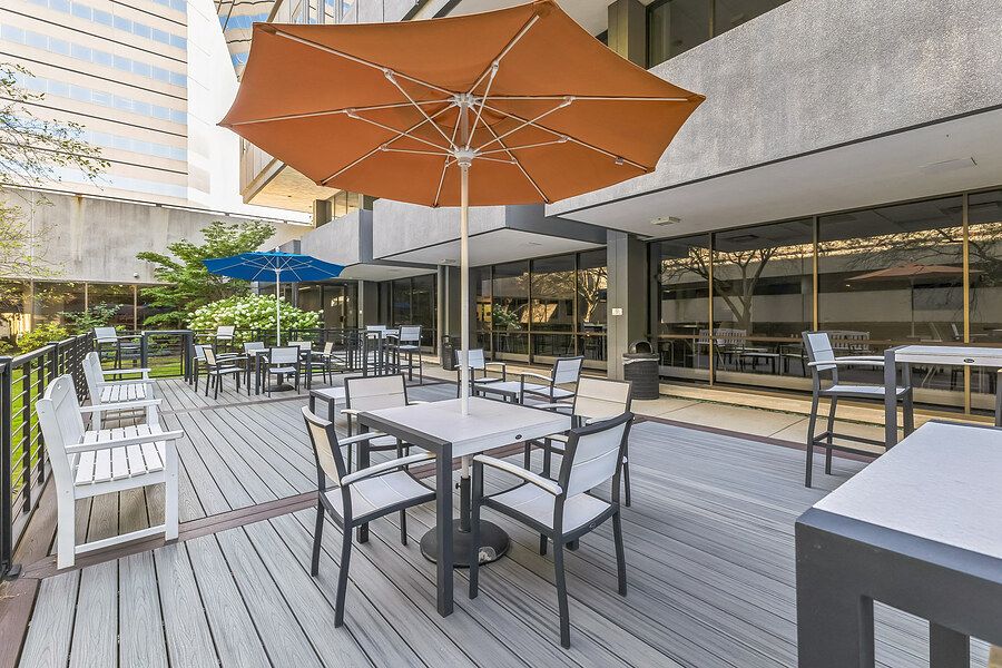 Modern office building outdoor patio with gray deck, dining tables, chairs, and orange market umbrella surrounded by greenery.