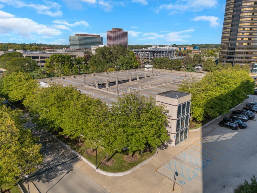 Aerial view of a multi-level parking garage surrounded by lush trees in an urban setting with office buildings in the background.