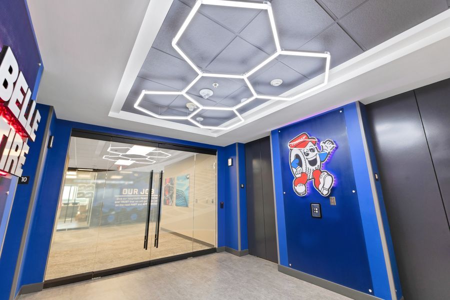 Modern office lobby with hexagonal ceiling lights, blue walls, branded signage, and a colorful mascot graphic near elevator doors.