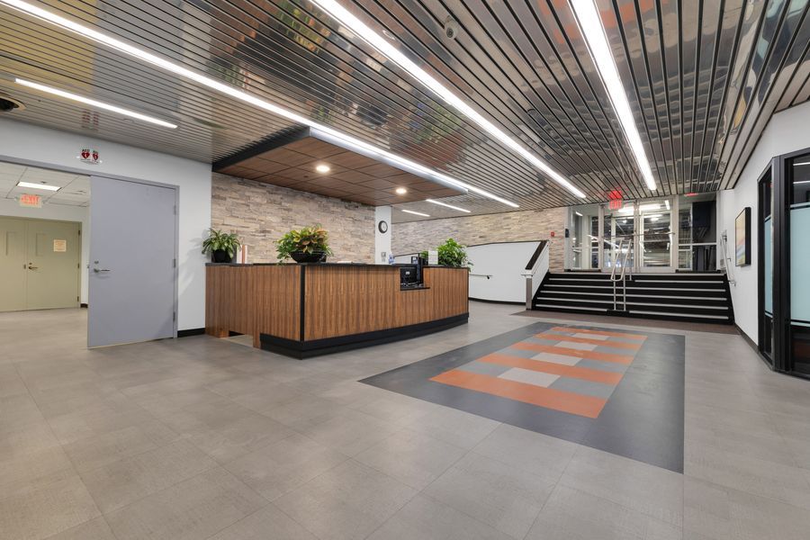 Modern office lobby with wooden reception desk, striped ceiling lights, orange accent floor tiles, and staircase in background.
