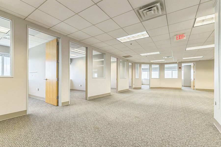 Spacious open-plan office interior with carpeted floors, drop ceiling, glass partition walls, and natural lighting.