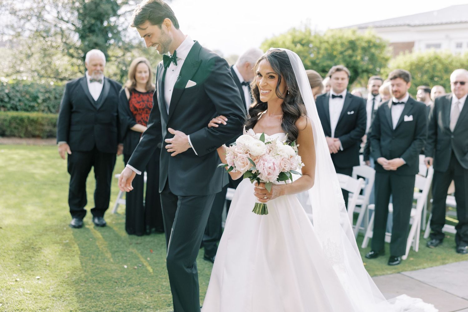 Bride in white ballgown holds pink bouquet walking down aisle with groom in dark tuxedo at outdoor wedding ceremony.