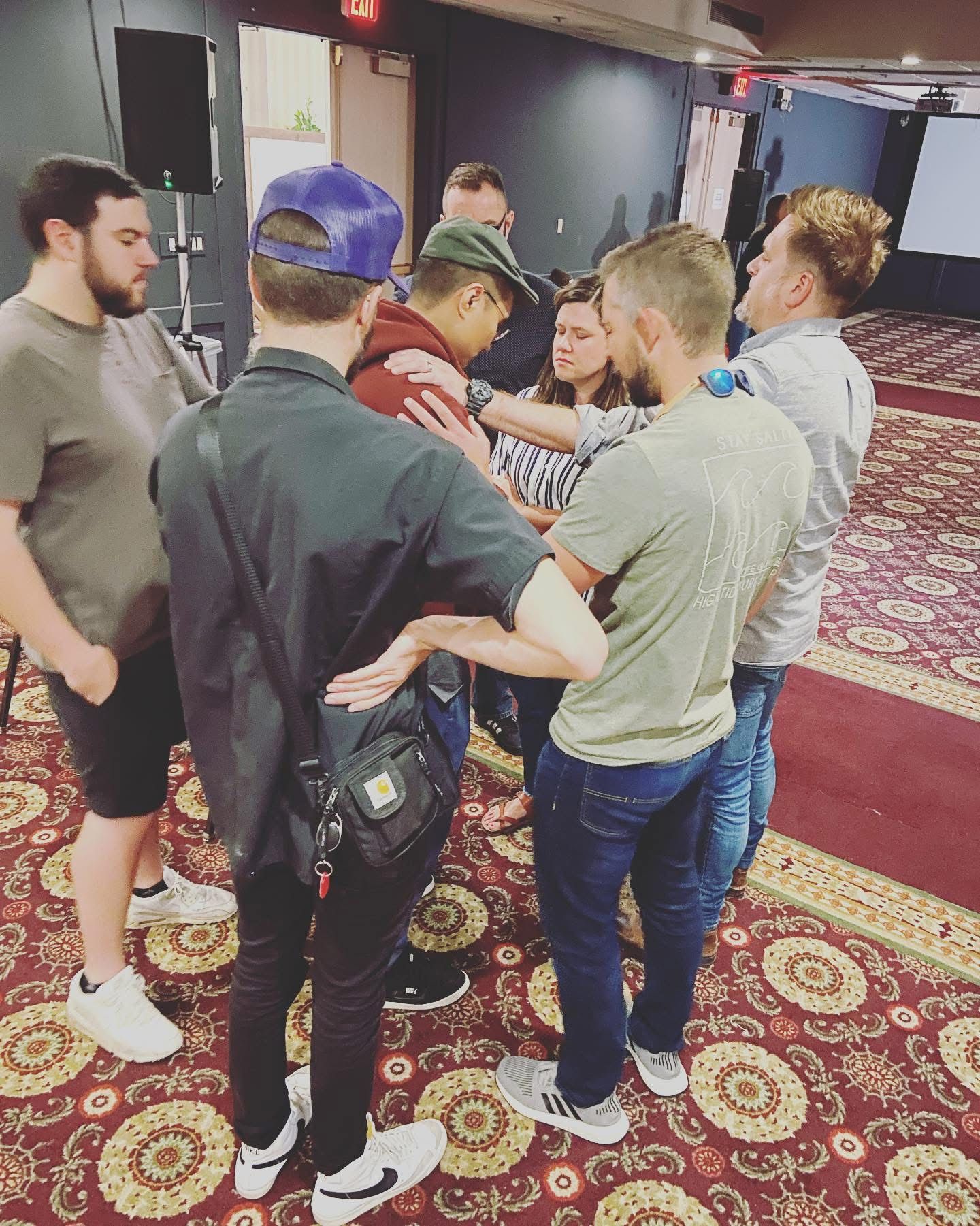 Group of young adults in a circle with hands on shoulders in a prayer huddle inside a venue with patterned carpet.