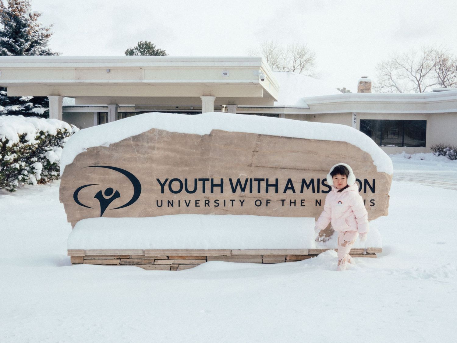 A snow-covered Youth With A Mission University of the Nations stone sign outside a building in winter.