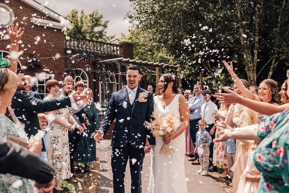 Bride and groom walk through confetti shower surrounded by cheering wedding guests outside a brick venue.