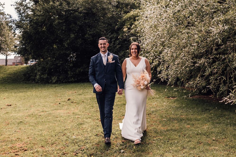Smiling bride and groom walking hand in hand on green grass with lush trees in background at outdoor wedding.