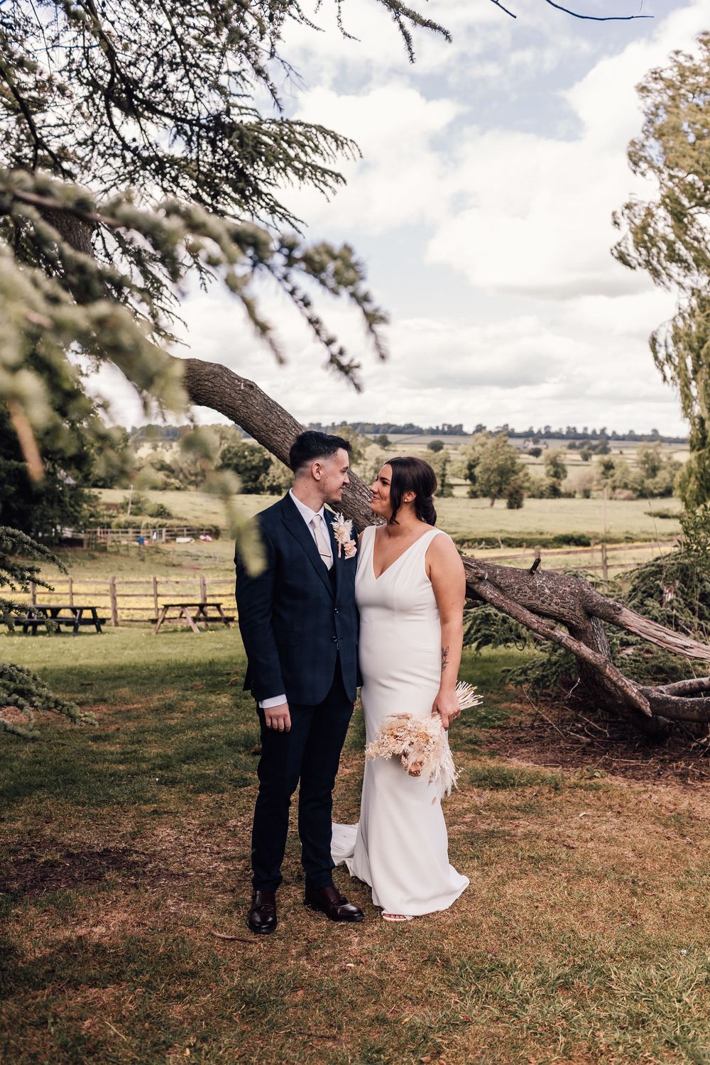 Bride and groom share a romantic moment under a tree in a lush countryside setting, bride holding a bouquet.