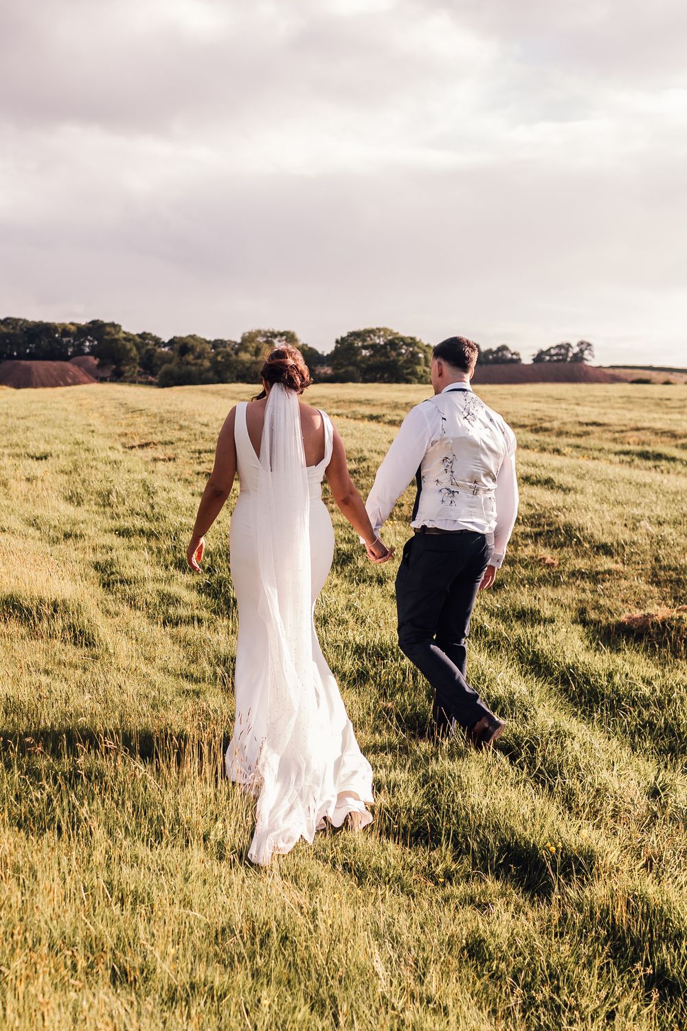 A bride and groom walk hand in hand through a lush green field on their wedding day.