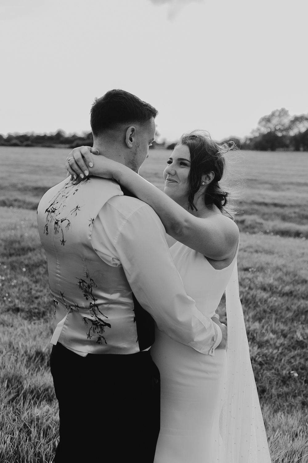 Black and white photo of a bride and groom embracing in a field, the groom wearing a patterned vest.