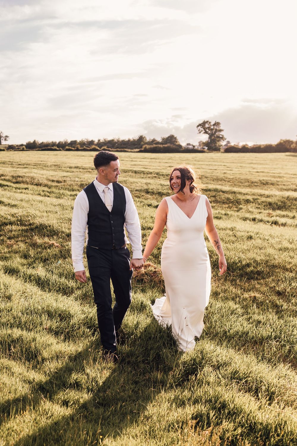 A smiling bride and groom hold hands walking through a sunlit green field on their wedding day.