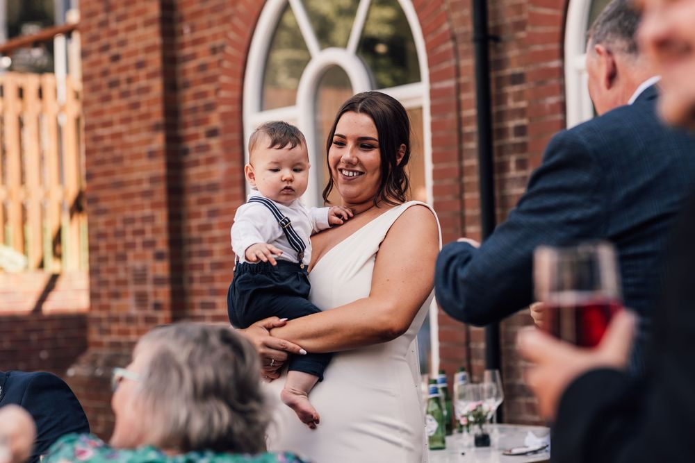 Bride in white dress holding baby at outdoor wedding reception near brick building.