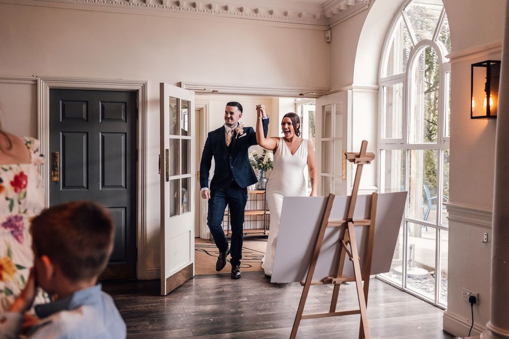 Bride and groom entering wedding reception hall, with easel displays and guests in elegant venue.