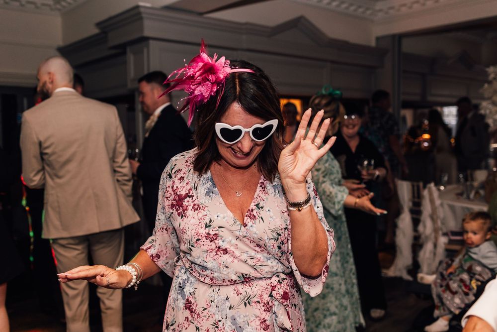 Woman in floral dress and white cat-eye sunglasses with pink fascinator dancing at a wedding reception.