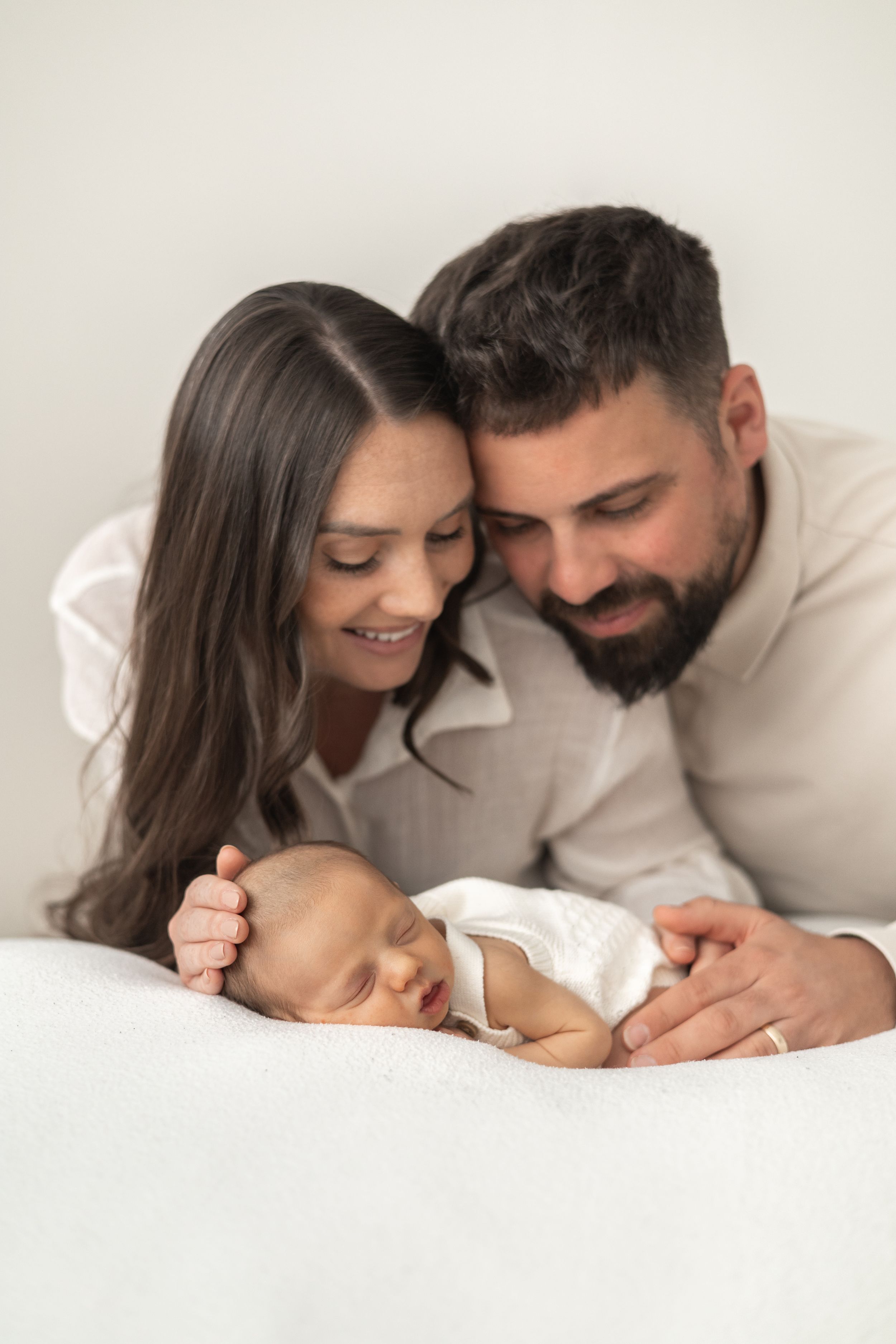 Happy parents smiling at their sleeping newborn baby lying on a white blanket in a natural newborn photoshoot Nottingham
