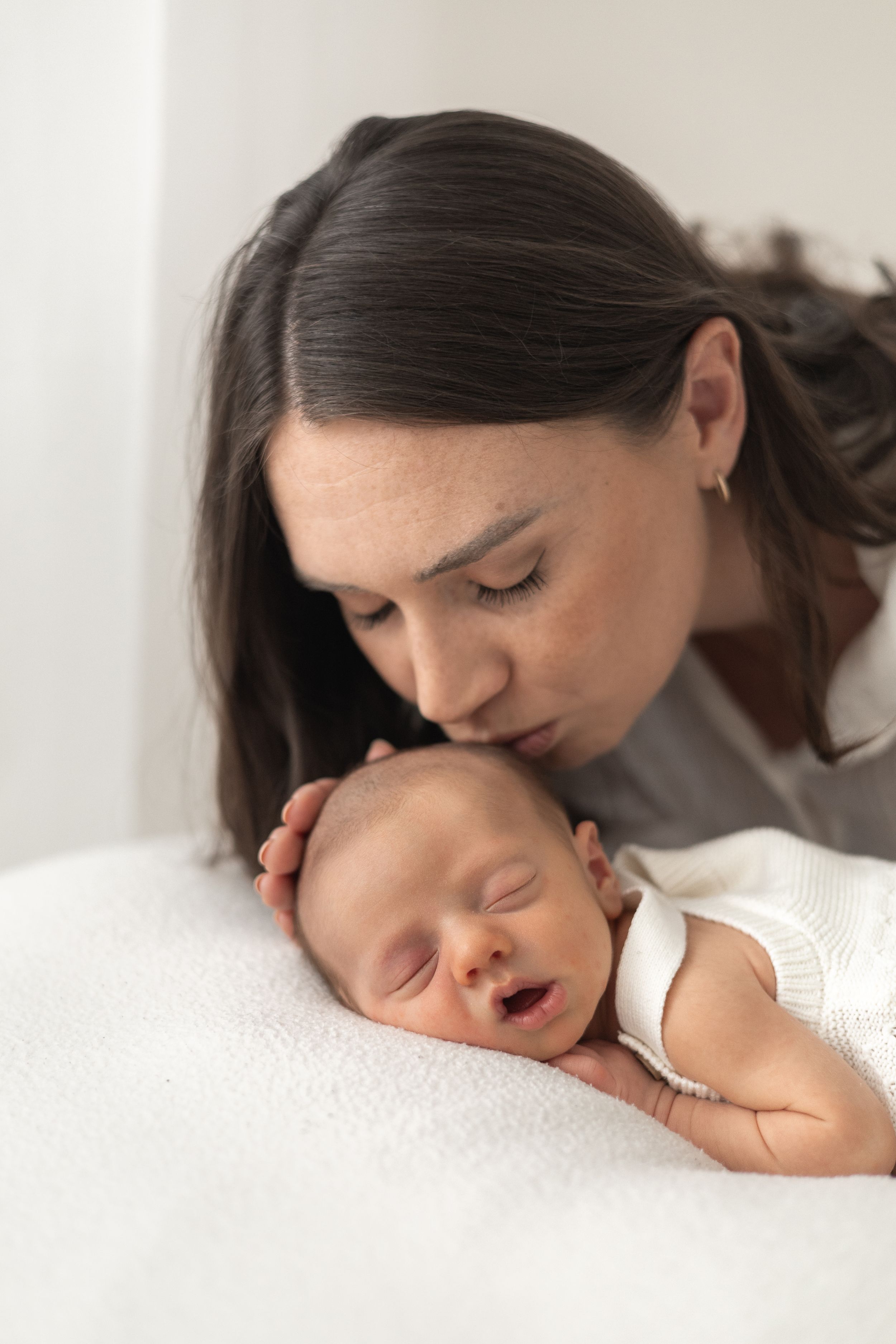 A mother tenderly kisses her sleeping newborn baby resting on a soft white blanket natural newborn photography Nottingham