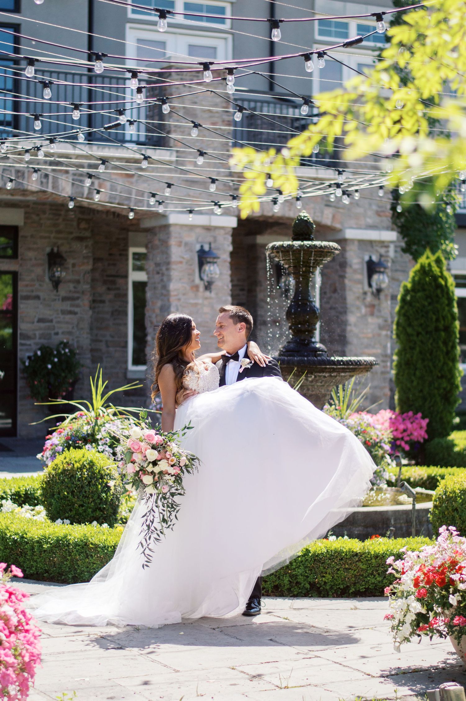 Groom lifts bride in white gown near garden fountain, string lights overhead, colorful flowers surrounding them.
