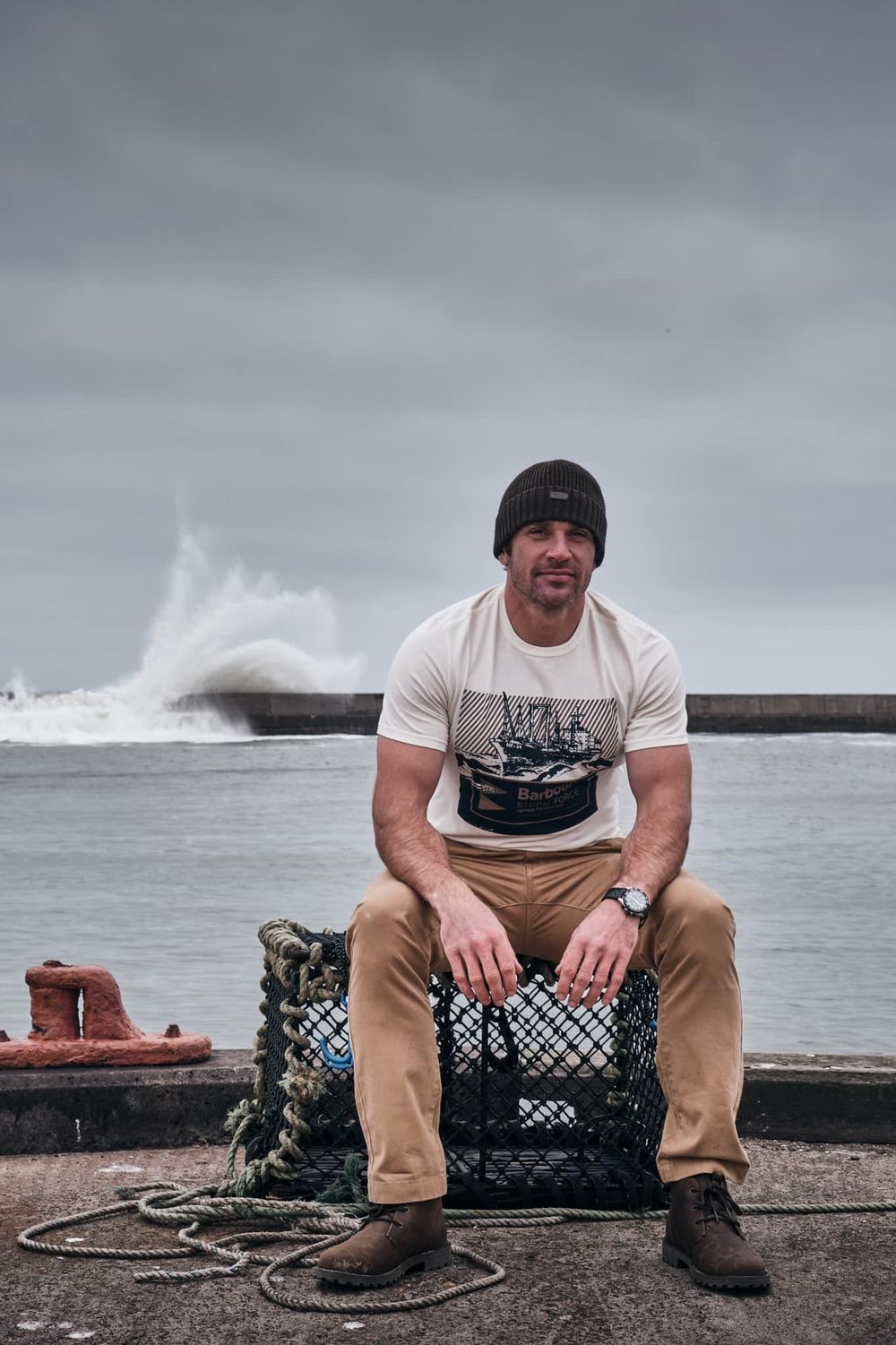 Muscular man in white graphic tee and beanie sits on lobster trap at stormy harbor with crashing waves behind him.