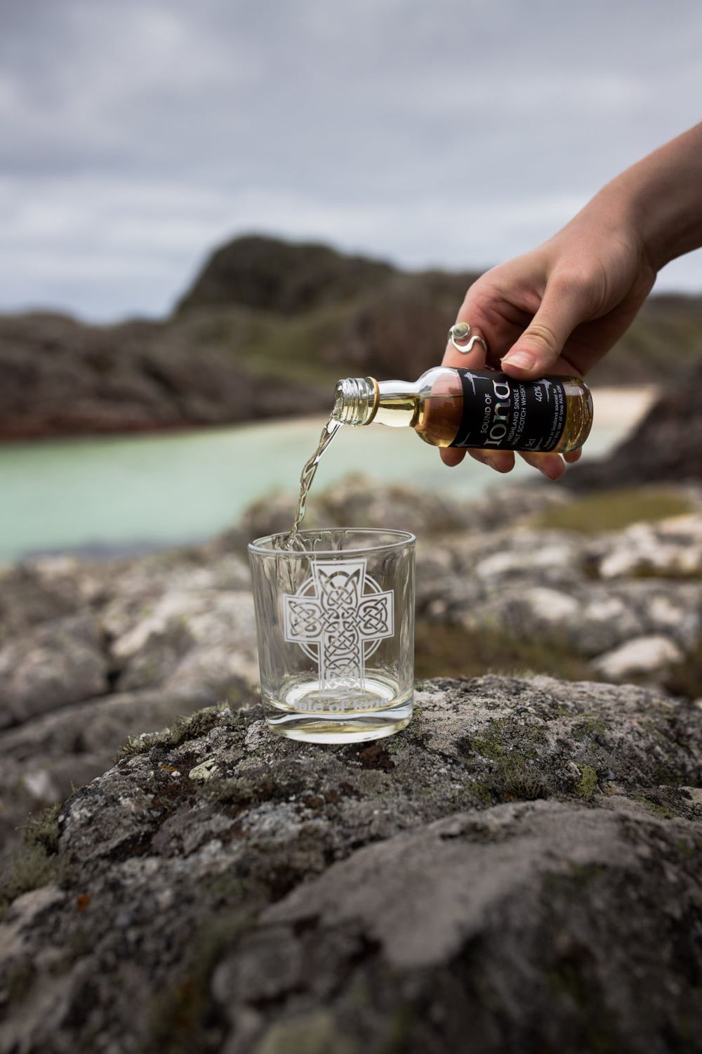 Hand pouring whisky from small bottle into Celtic cross engraved glass on rocky Scottish coastal landscape.
