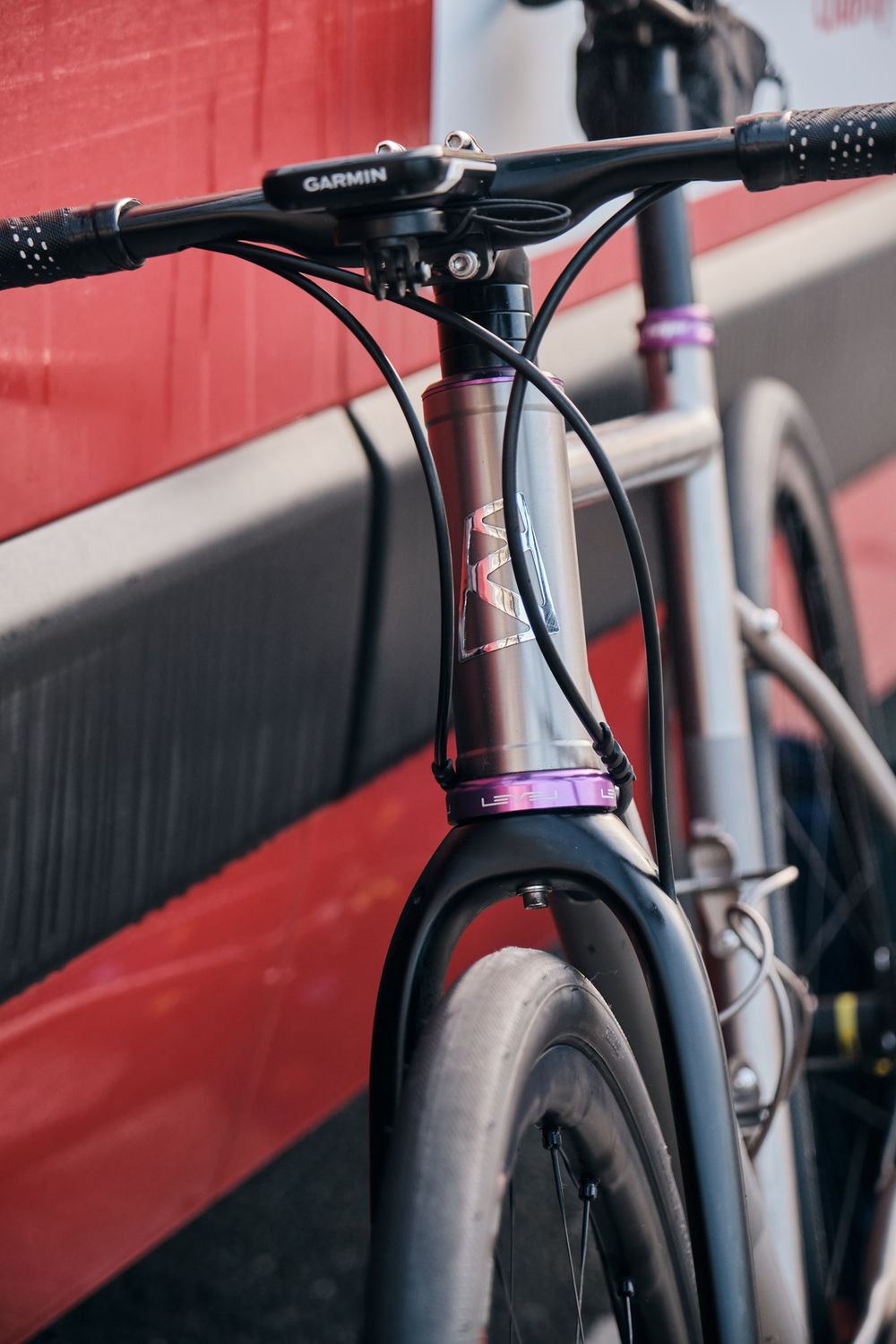 Close-up of a road bike handlebar with Garmin mount, purple headset spacer, and carbon fork leaning against a red wall.