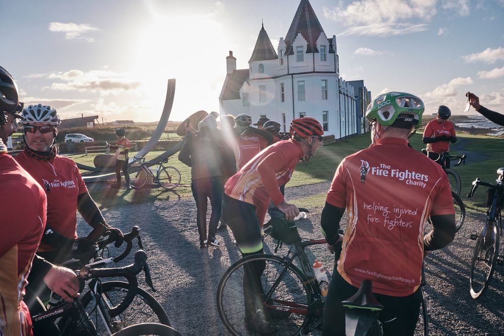 Cyclists in red jerseys gather near a white church on a sunny day during a charity bike ride event.
