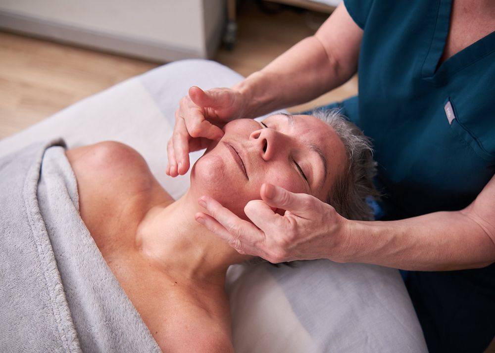 A therapist performs a relaxing facial massage on a senior woman lying on a spa treatment table.