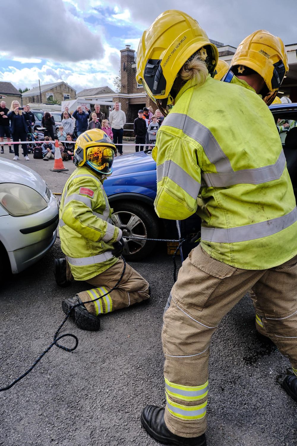 Firefighters in yellow gear work on a crashed car during a rescue demonstration while spectators watch in the background.