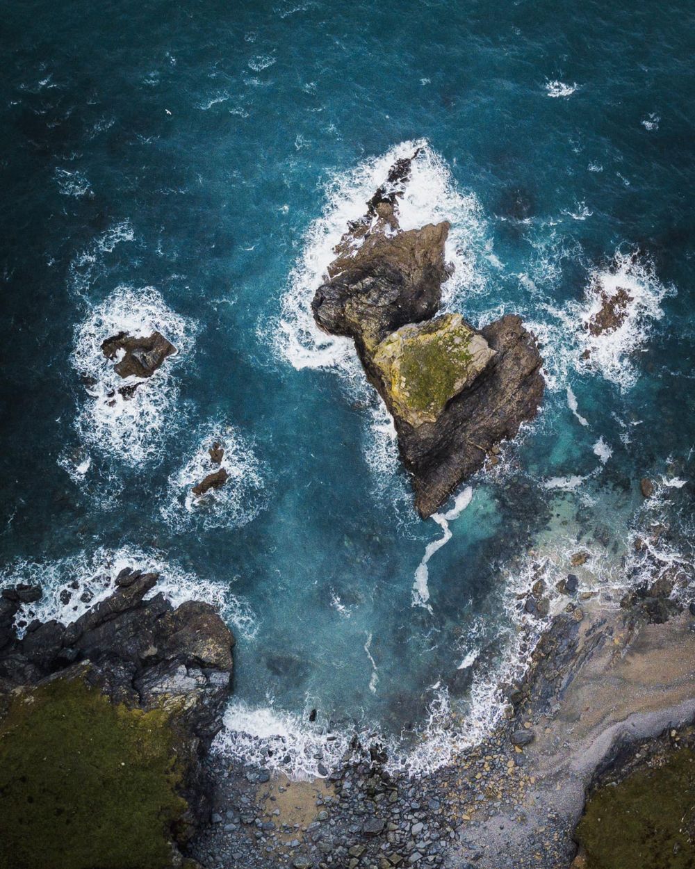 Aerial view of a rocky outcrop surrounded by turquoise ocean waves crashing against the rugged coastline.