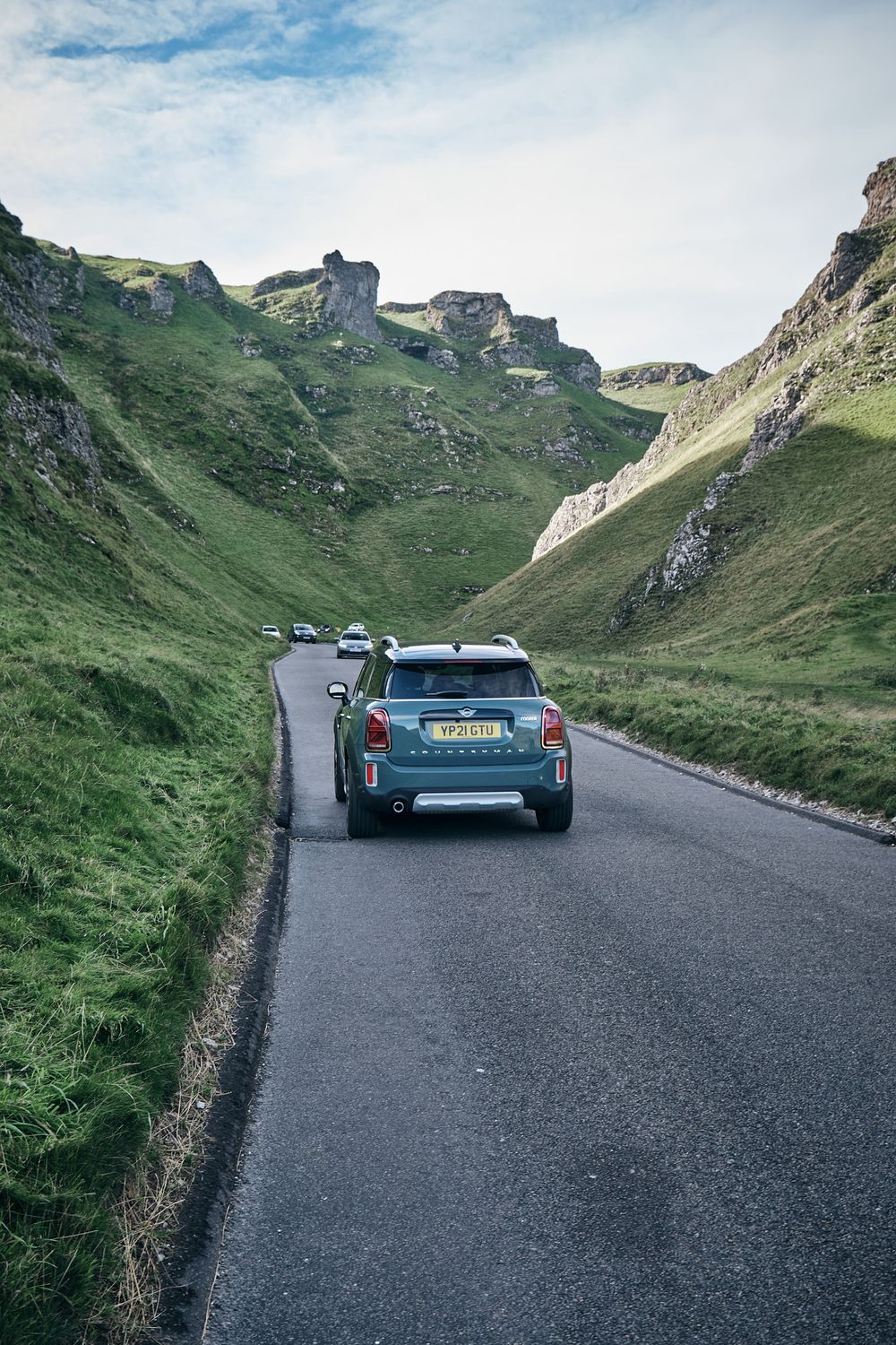 Cars driving on a narrow road through a lush green valley with rocky hills under a partly cloudy sky.