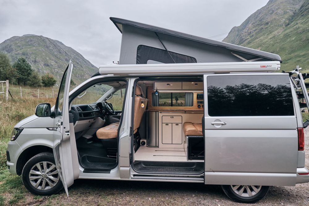 Silver VW California campervan with pop-up roof open, showing interior conversion, parked in Scottish Highland mountains.