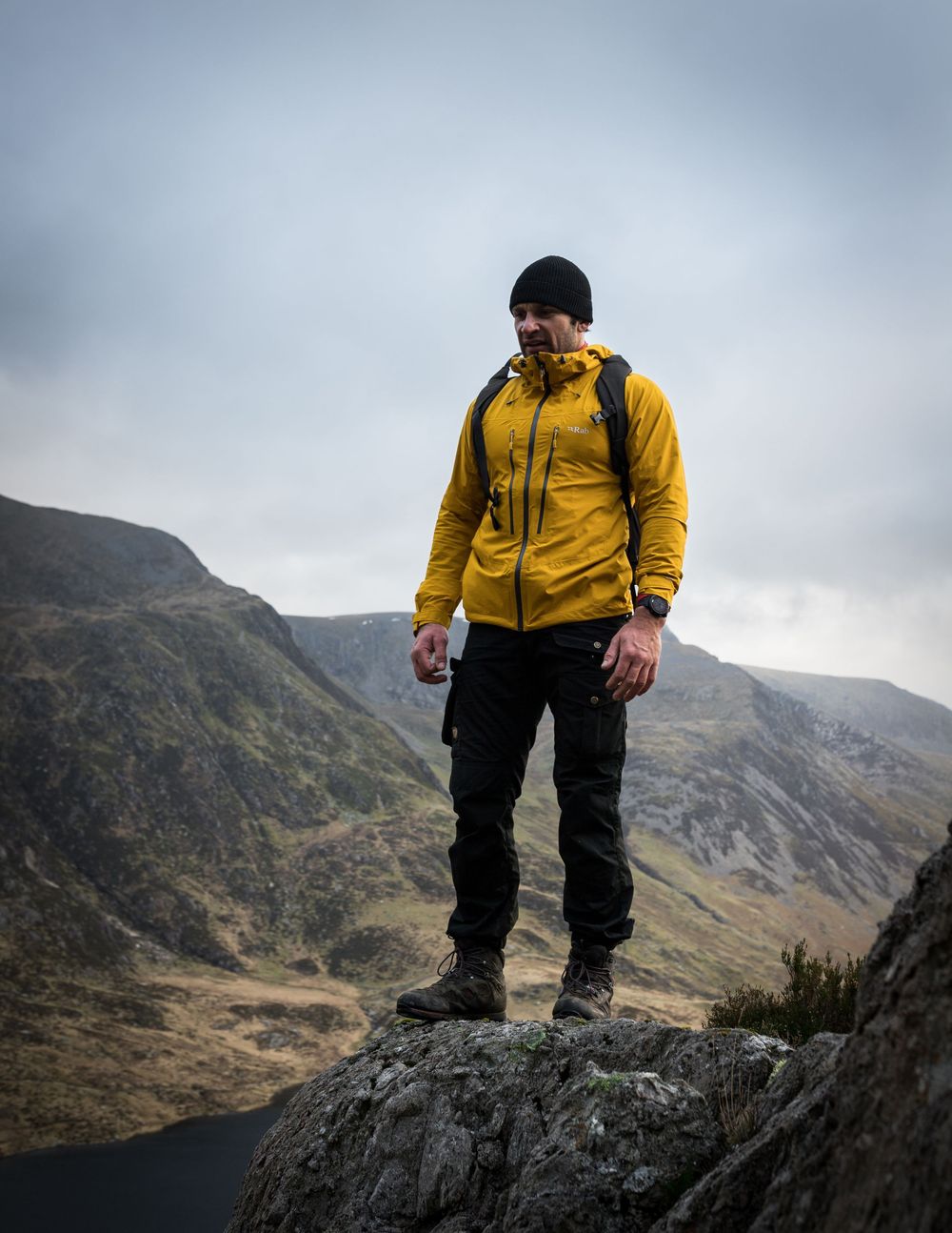 Hiker in yellow jacket standing on rocky summit with dramatic mountain valley backdrop.