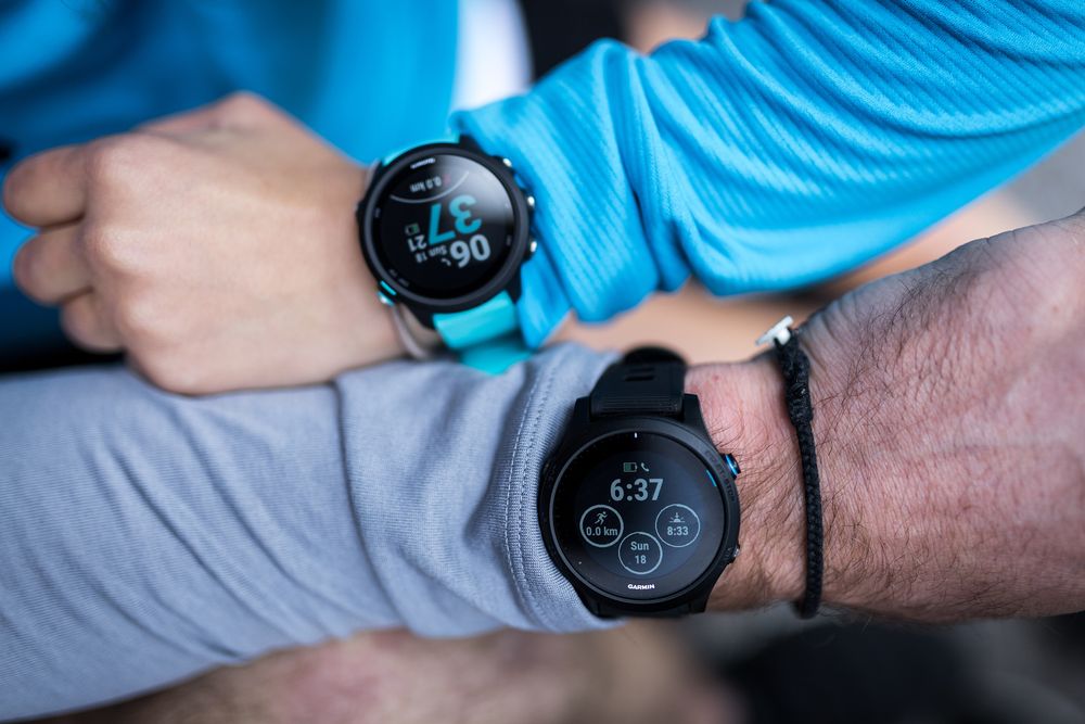 Two Garmin smartwatches displayed on wrists, showing fitness tracking data including heart rate and time.