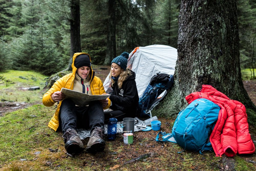 Two hikers in yellow jackets review a map while camping in a misty forest with backpacks and gear nearby.